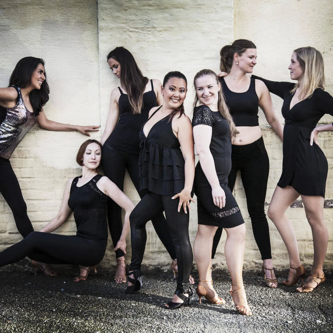 A group of dancers in black posing against a white brick wall